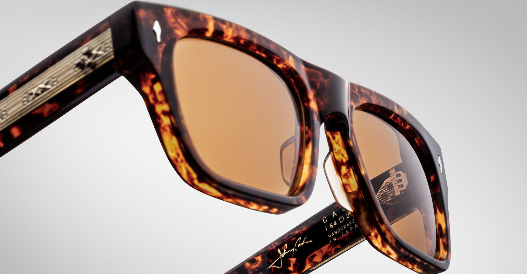 Close-up of Jacques Marie Mage Cash sunglasses with tortoiseshell frame and amber lenses, shown against a plain background. Features gold detailing on the arms plus visible branding and signatures inside the arms.