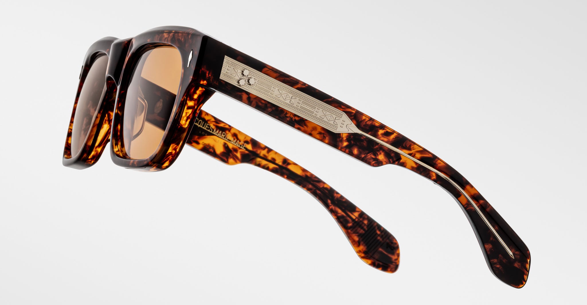 A close-up of Jacques Marie Mage Cash sunglasses featuring tortoiseshell-patterned rectangular frames, brown lenses, and arms adorned with a metallic plaque engraved with text and a small emblem, set against a plain white background.