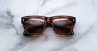 A pair of Jacques Marie Mage Cash rectangular tortoiseshell sunglasses with brown lenses rests on a white marble surface, featuring some text and reflections visible on the lenses.