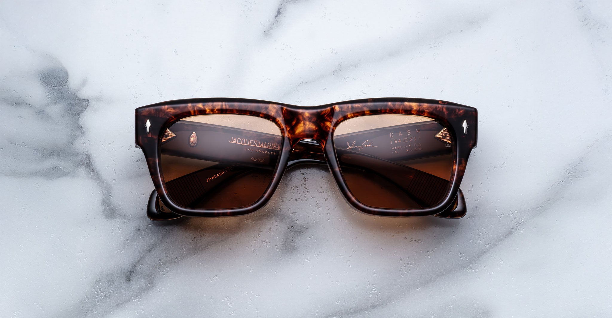 A pair of Jacques Marie Mage Cash rectangular tortoiseshell sunglasses with brown lenses rests on a white marble surface, featuring some text and reflections visible on the lenses.