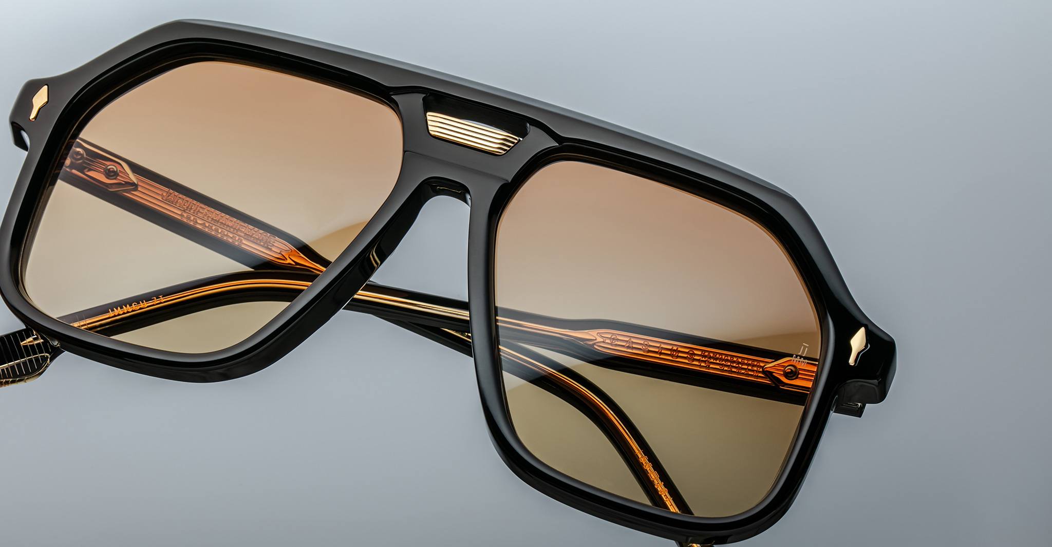 Close-up of Jacques Marie Mage Casius sunglasses with black angular frames, gold temple detailing, and brown tinted lenses, displayed on a light grey background.