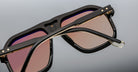 Close-up of Jacques Marie Mage Casius sunglasses with thick black frames, pink-tinted lenses, gold detailing, and small star-shaped accents on the folded hinges.