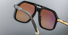 Close-up of Jacques Marie Mage Casius sunglasses featuring black and gold frames with brown-tinted lenses, set against a neutral gray background.