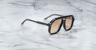 The Jacques Marie Mage Casius black sunglasses with brown-tinted lenses rest on a white marble surface with gray veining.
