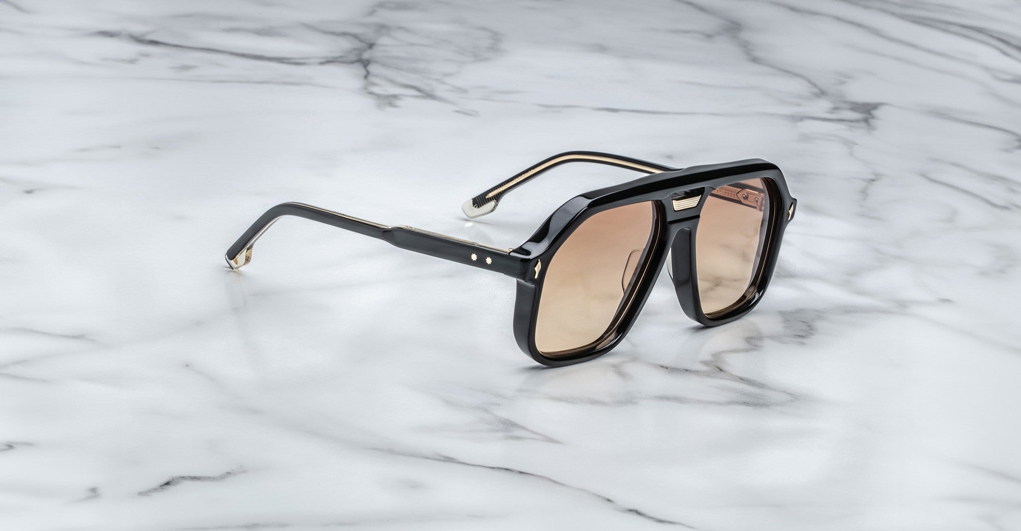 The Jacques Marie Mage Casius black sunglasses with brown-tinted lenses rest on a white marble surface with gray veining.