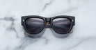 A pair of Jacques Marie Mage Chelsea brown, thick-framed sunglasses with purple lenses rests on a white marble surface with grey veins, with faint brand details visible on the inside of the left lens.