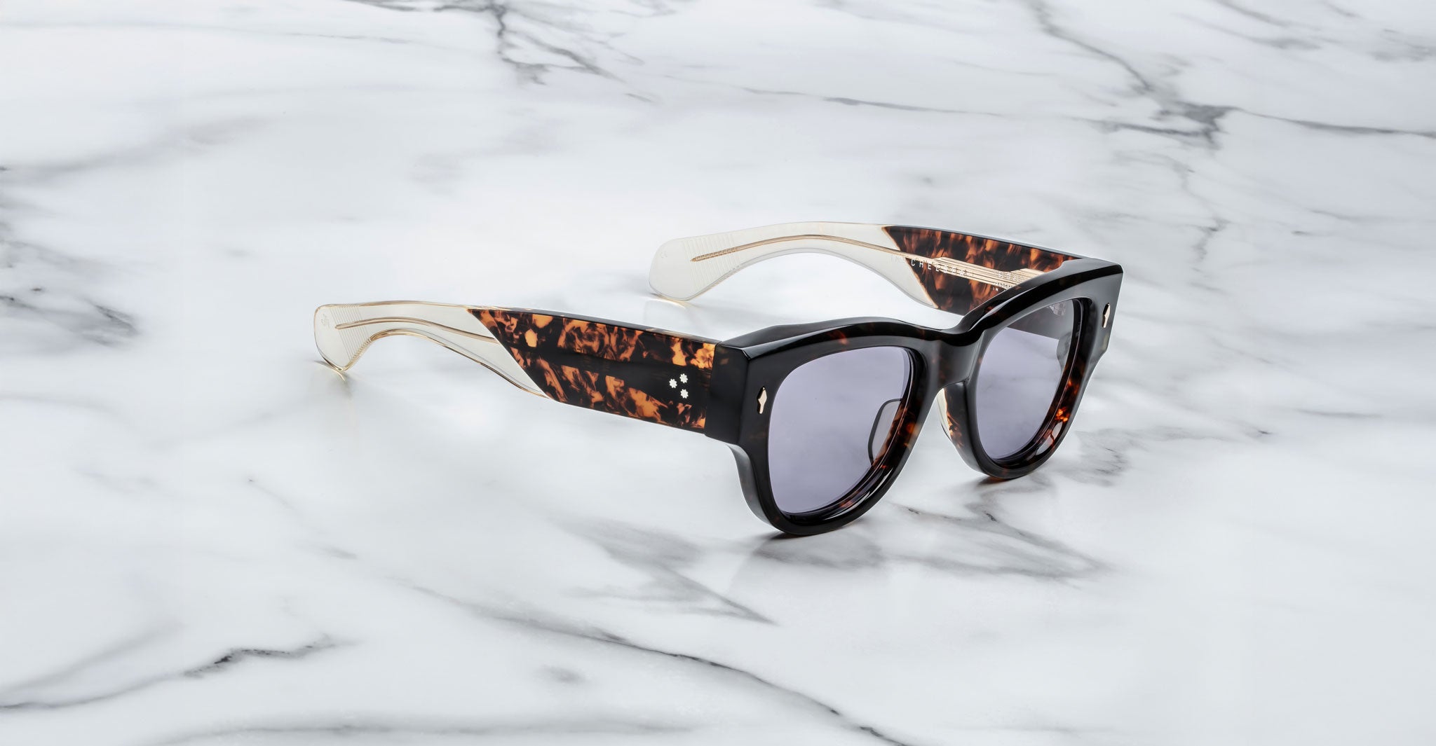 A pair of Jacques Marie Mage Chelsea brown, thick-framed sunglasses with purple lenses sits on a white marble surface with grey veins