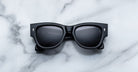 A pair of Jacques Marie Mage Chelsea black, thick-framed sunglasses with grey lenses rests on a white marble surface with grey veins, with faint brand details visible on the inside of the left lens.