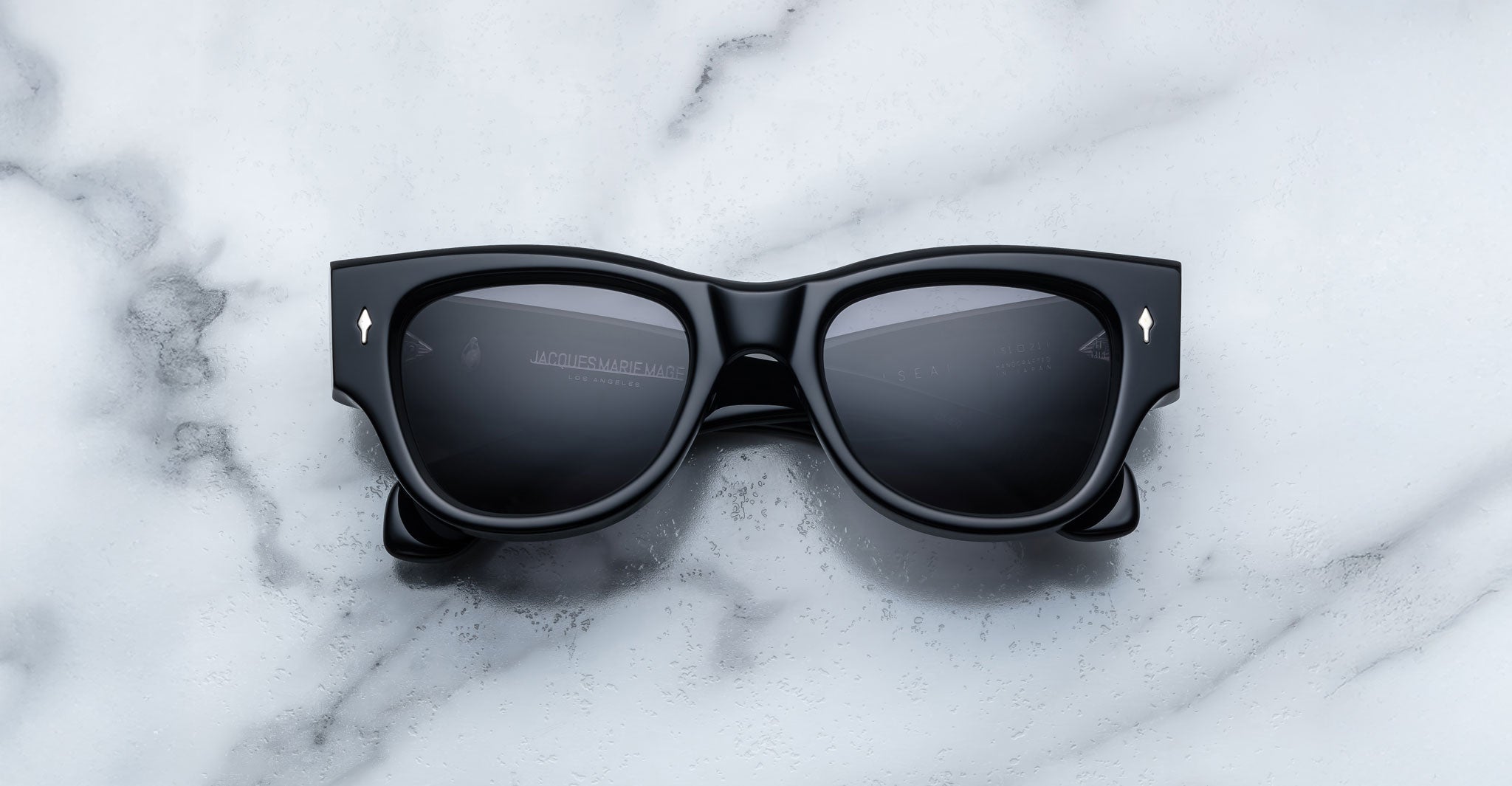 A pair of Jacques Marie Mage Chelsea black, thick-framed sunglasses with grey lenses rests on a white marble surface with grey veins, with faint brand details visible on the inside of the left lens.
