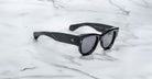 A pair of Jacques Marie Mage Chelsea black sunglasses with thick frames and dark lenses rests on a white marble surface, featuring small metal dots near the hinges.