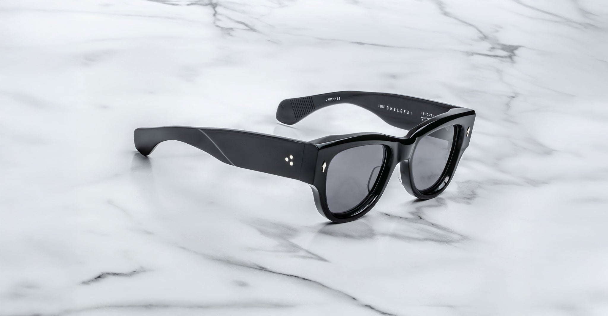 A pair of Jacques Marie Mage Chelsea black sunglasses with thick frames and dark lenses rests on a white marble surface, featuring small metal dots near the hinges.