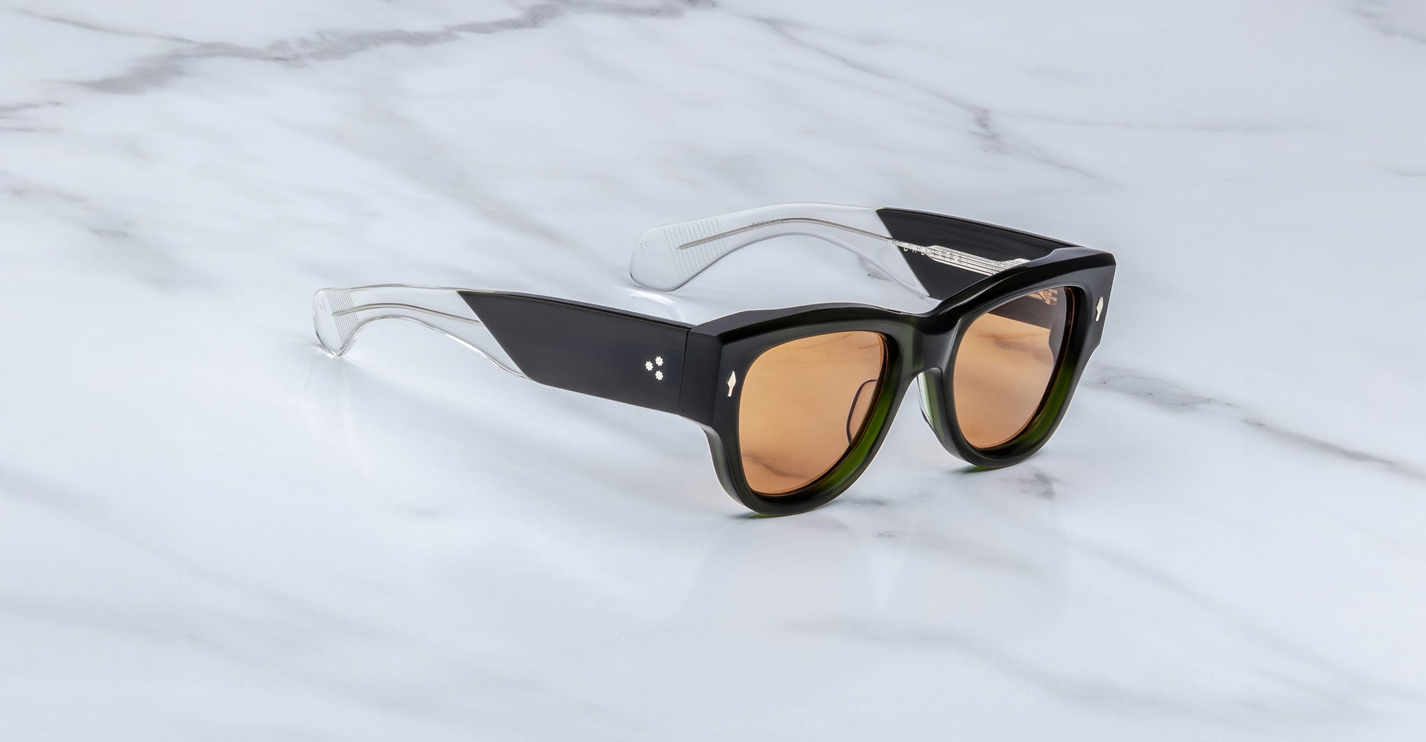 The Jacques Marie Mage Chelsea sunglasses, featuring thick black and translucent frames with brown-tinted lenses, rest on a white marble surface.