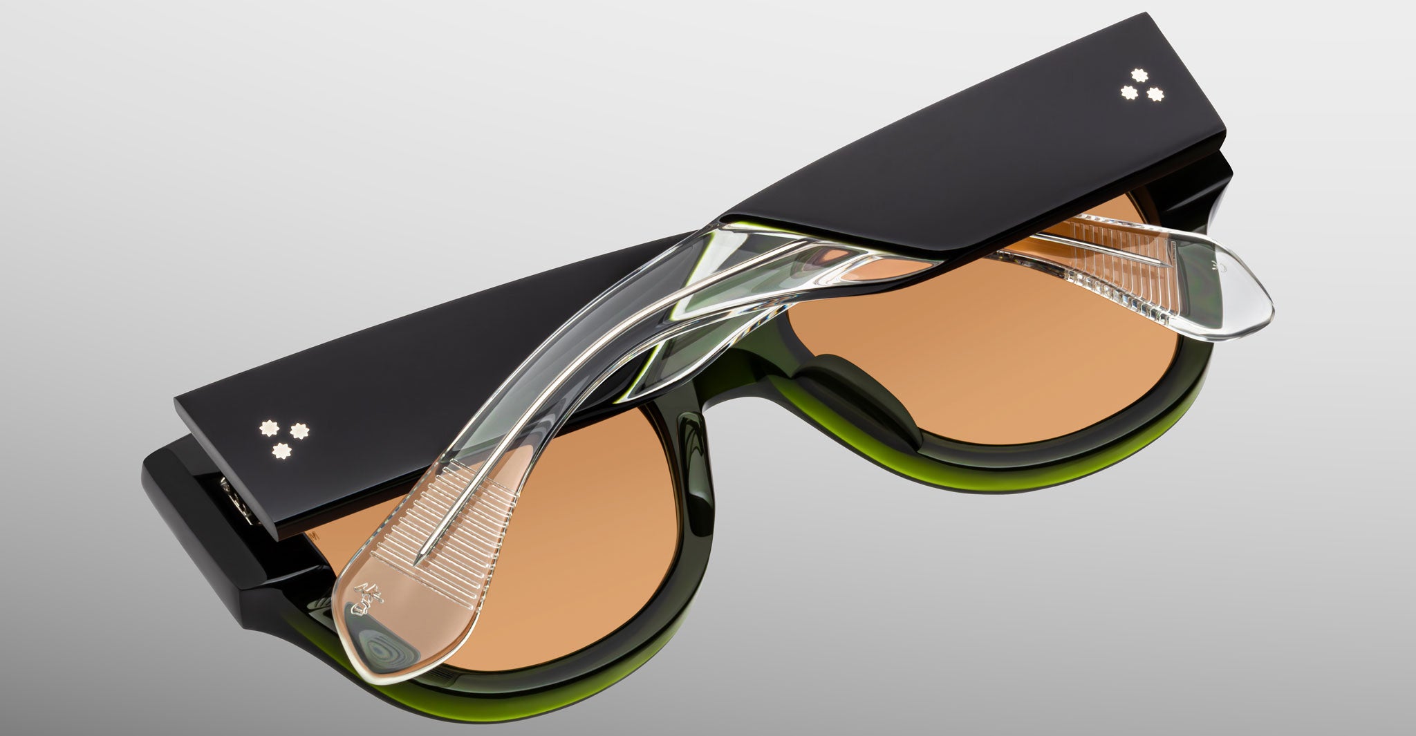The Jacques Marie Mage Chelsea sunglasses feature thick black frames, light brown lenses, and transparent textured arms with crossed temples, shown against a neutral gradient background.