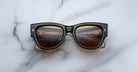 Jacques Marie Mage Chelsea black-framed sunglasses with dark lenses sit on a white marble surface, their lenses reflecting a warm, curved interior with ceiling lights and the words ACID CINNAMON ACID.
