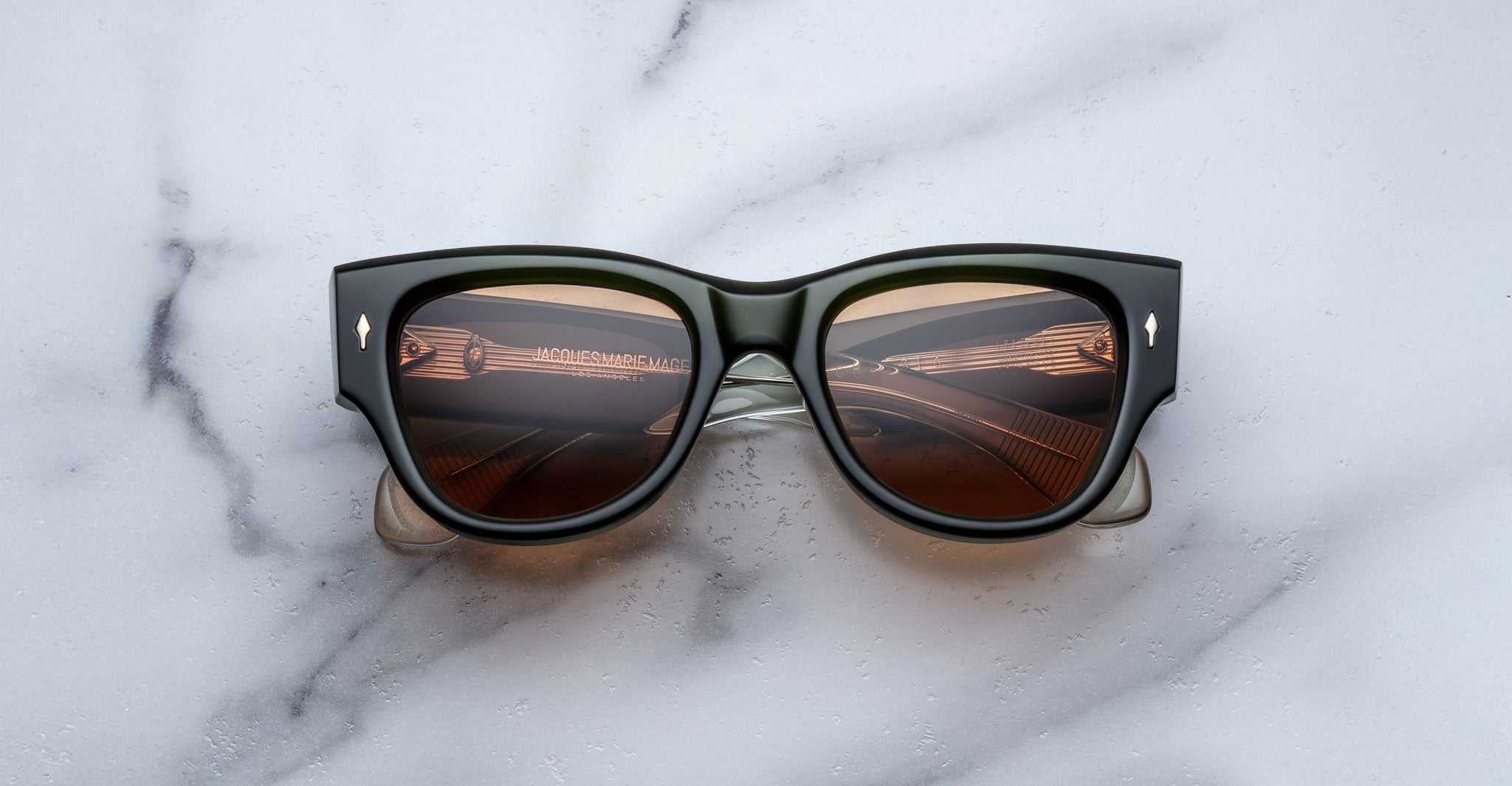 Jacques Marie Mage Chelsea black-framed sunglasses with dark lenses sit on a white marble surface, their lenses reflecting a warm, curved interior with ceiling lights and the words ACID CINNAMON ACID.