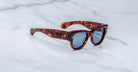The Jacques Marie Mage Chelsea sunglasses, featuring thick dark tortoiseshell frames and dark tinted lenses, rest on a smooth white marble surface.