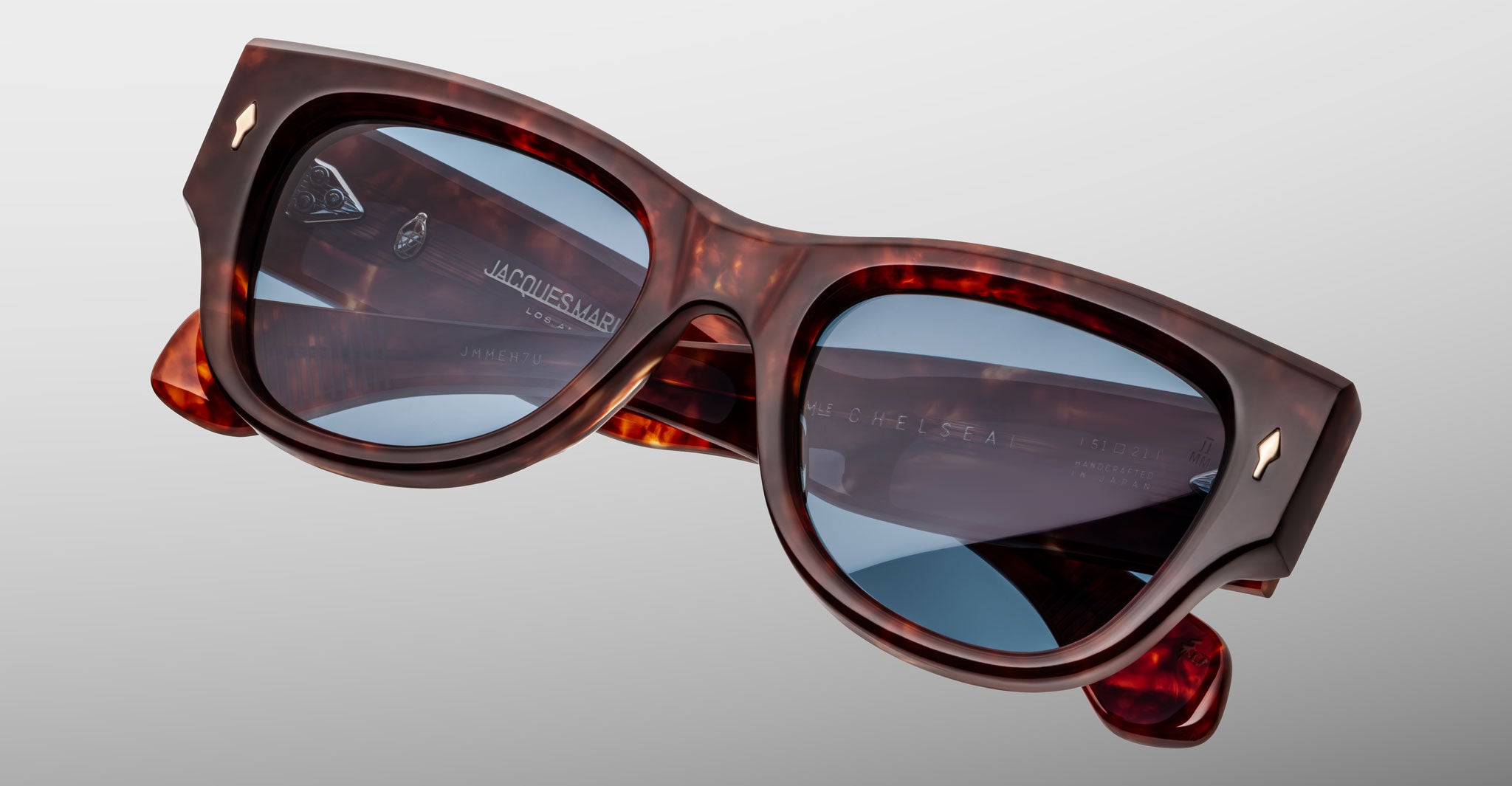 A close-up of Jacques Marie Mage Chelsea sunglasses featuring thick tortoiseshell frames and blue-tinted lenses against a plain light gray background.