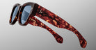 Close-up of Jacques Marie Mage Chelsea sunglasses featuring thick red and brown tortoiseshell frames, blue-tinted lenses, and two small star-like dots on the temple, set against a neutral light gray background.