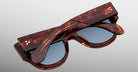 The Jacques Marie Mage Chelsea tortoiseshell sunglasses with blue lenses are shown folded closed, arms crossed over the front, on a plain light background.