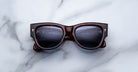 A pair of Jacques Marie Mage Chelsea tortoiseshell sunglasses with black lenses rests on a white marble surface, reflecting a faint image of a computer screen and keyboard.