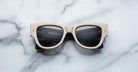 A pair of Jacques Marie Mage Chelsea beige, thick-framed sunglasses with dark lenses rests on a white marble surface with grey veins, with faint brand details visible on the inside of the left lens.