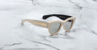 A pair of Jacques Marie Mage Chelsea beige, thick-framed sunglasses with dark lenses sits on a white marble surface with grey veins, with faint brand details visible on the inside of the left lens.