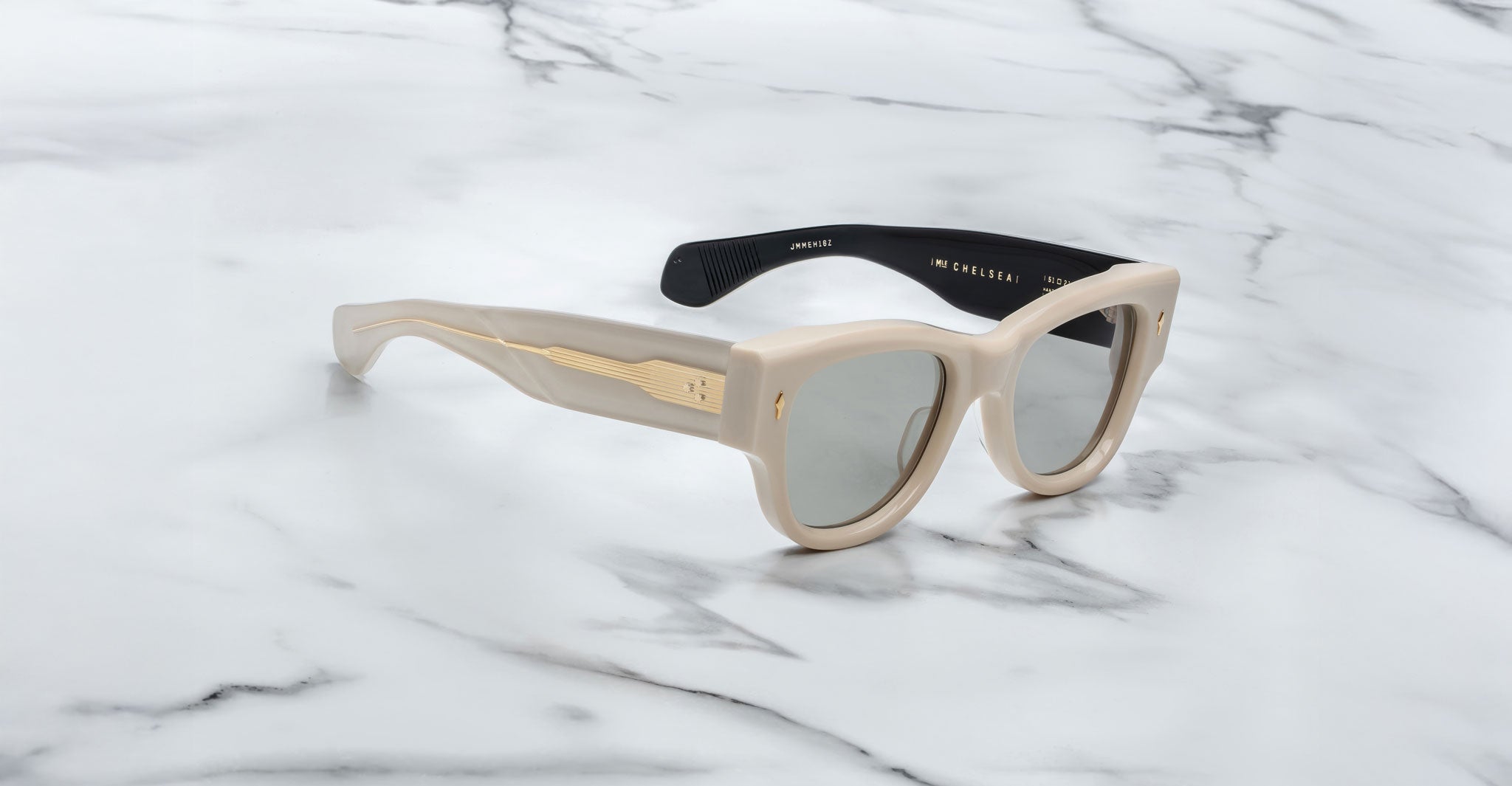 A pair of Jacques Marie Mage Chelsea beige, thick-framed sunglasses with dark lenses sits on a white marble surface with grey veins, with faint brand details visible on the inside of the left lens.