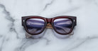 A pair of Jacques Marie Mage Chelsea cherry red, thick-framed sunglasses with light purple lenses rests on a white marble surface with grey veins, with faint brand details visible on the inside of the left lens.