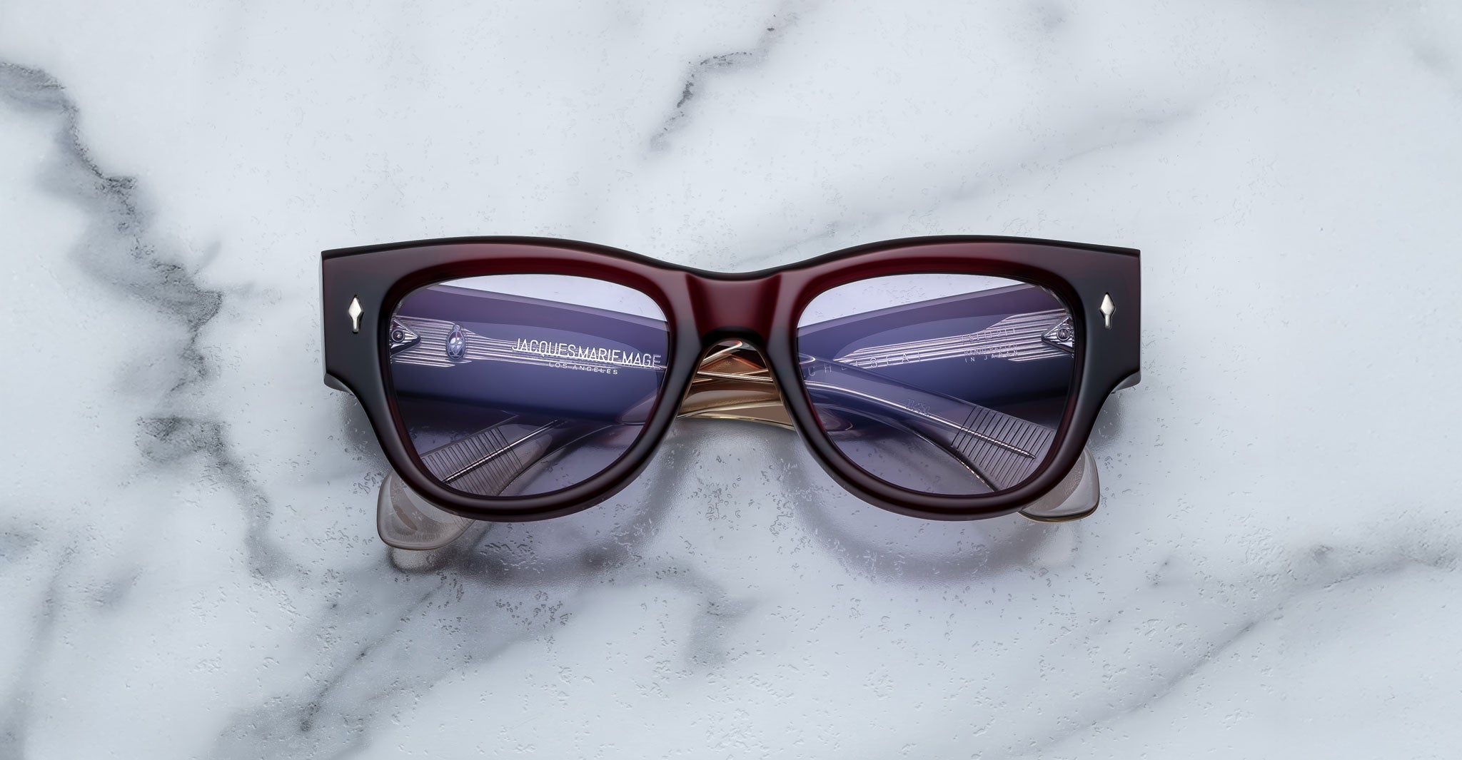 A pair of Jacques Marie Mage Chelsea cherry red, thick-framed sunglasses with light purple lenses rests on a white marble surface with grey veins, with faint brand details visible on the inside of the left lens.