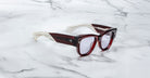 A pair of Jacques Marie Mage Chelsea cherry red, thick-framed sunglasses with light purple lenses sits on a white marble surface with grey veins