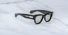 A pair of Jacques Marie Mage Chelsea sunglasses with black frames, dark green arms, and silver detailing is displayed on a white marble surface. The inner left arm features the text AVIONIC.