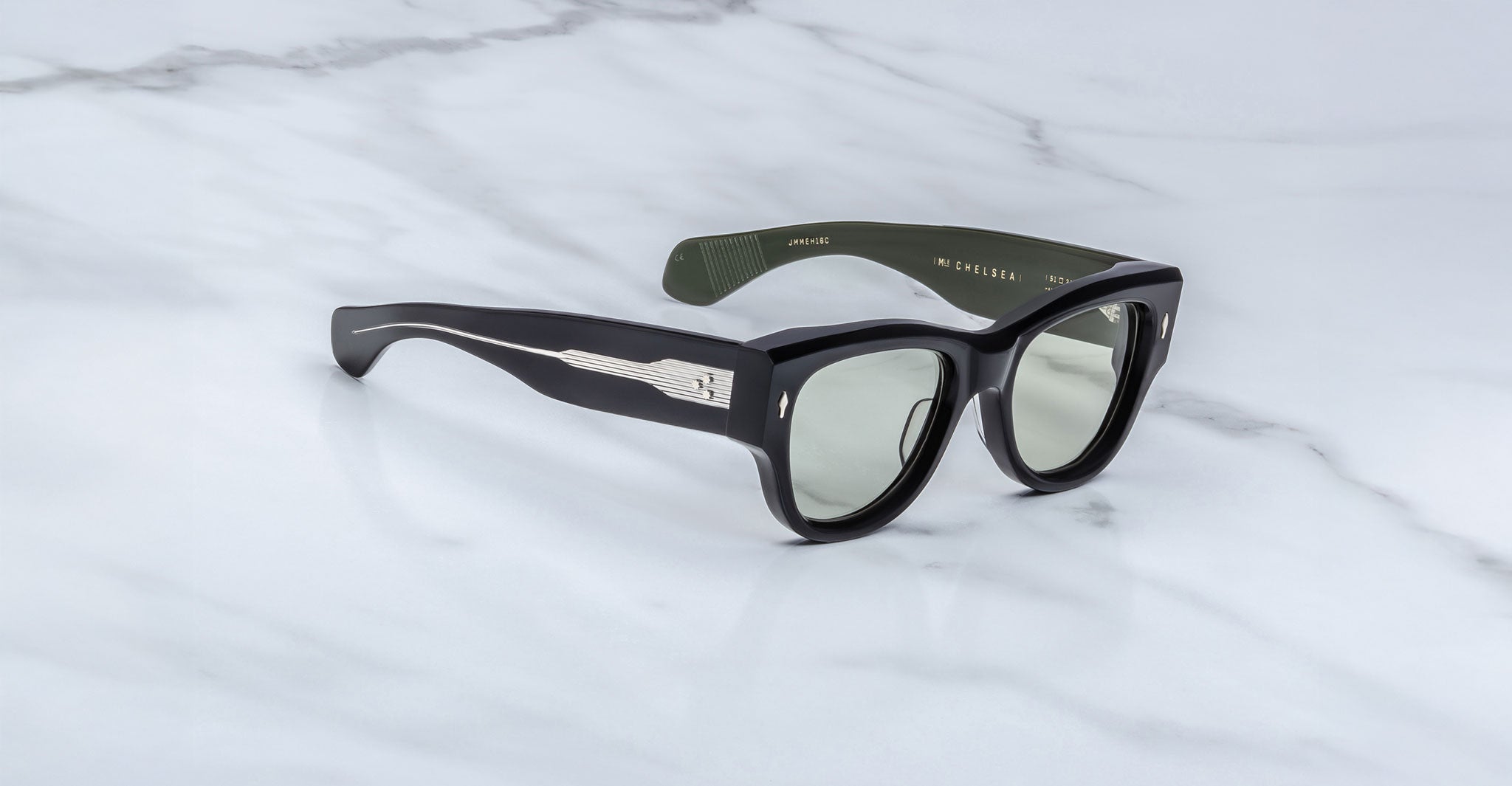A pair of Jacques Marie Mage Chelsea sunglasses with black frames, dark green arms, and silver detailing is displayed on a white marble surface. The inner left arm features the text AVIONIC.