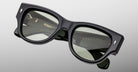 The Jacques Marie Mage Chelsea is a pair of dark, rectangular sunglasses with thick black frames and green-tinted lenses, shown close-up on a light gray background. The inner arms feature engraved gold branding and details.