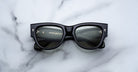 A pair of Jacques Marie Mage Chelsea black rectangular sunglasses with thick frames rests on a white marble surface, their lenses reflecting text and markings that indicate branding or manufacturer details inside the frame.