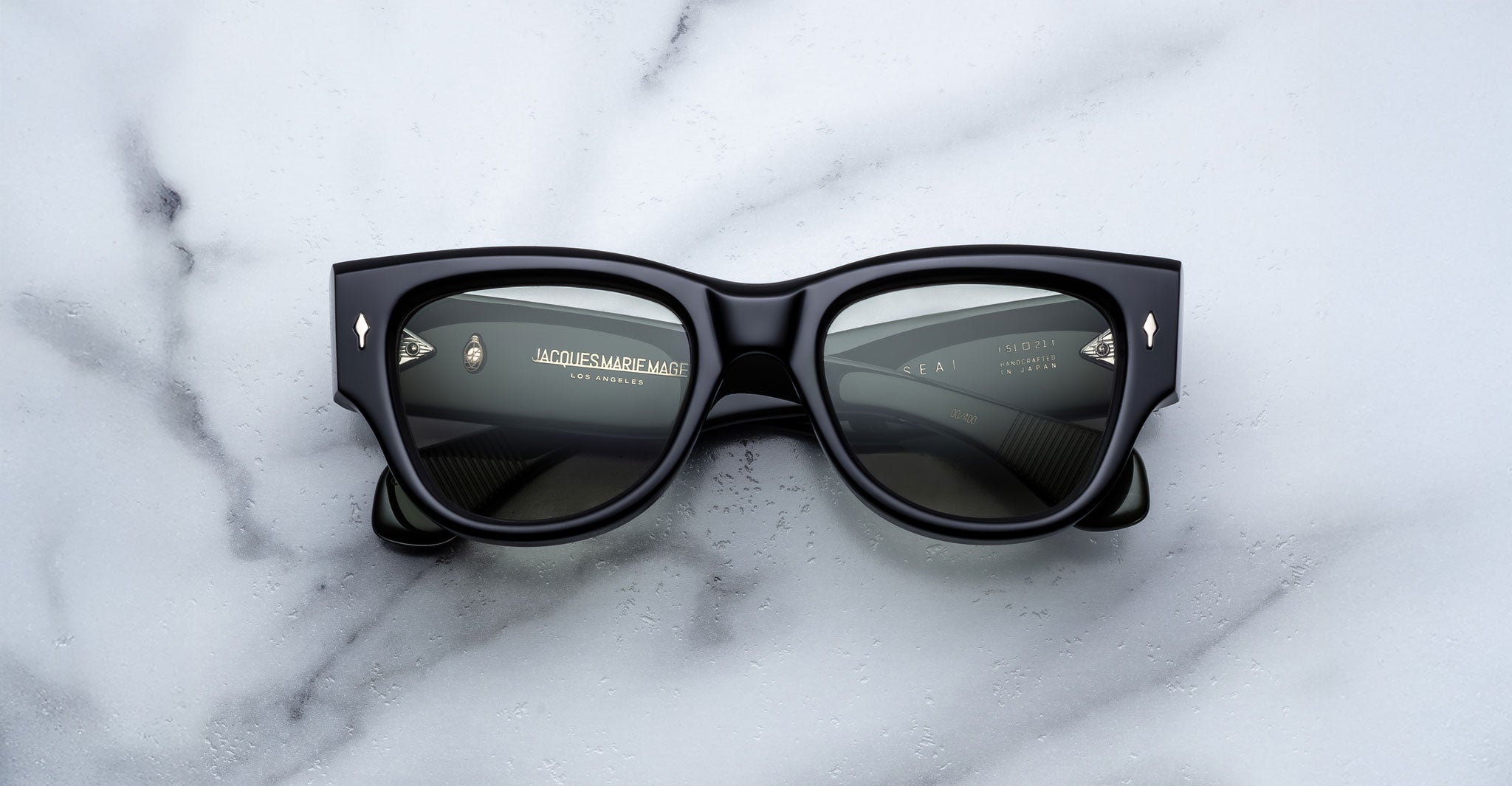 A pair of Jacques Marie Mage Chelsea black rectangular sunglasses with thick frames rests on a white marble surface, their lenses reflecting text and markings that indicate branding or manufacturer details inside the frame.