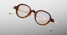 The Jacques Marie Mage Clark eyeglasses by Jacques Marie Mage feature brown tortoiseshell round frames with gold metal temples, handcrafted in Japan and shown on a light gray background.