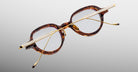 The Jacques Marie Mage Clark eyeglasses feature round tortoiseshell frames and gold metal arms, handcrafted in Japan with midcentury inspiration, displayed top-down on a light gray background.