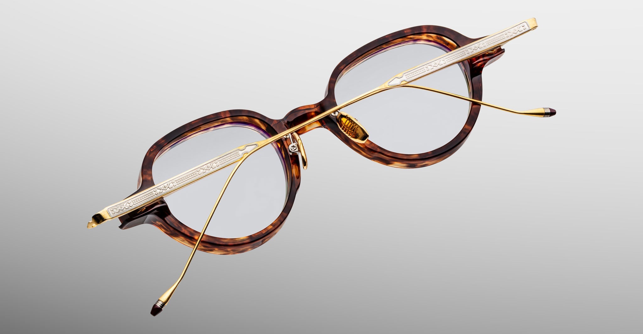 The Jacques Marie Mage Clark eyeglasses feature round tortoiseshell frames and gold metal arms, handcrafted in Japan with midcentury inspiration, displayed top-down on a light gray background.