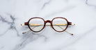 Jacques Marie Mage Clark eyeglasses by Jacques Marie Mage, featuring round tortoiseshell frames and gold titanium temples handcrafted in Japan, are displayed on a white marble surface.