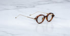 The Jacques Marie Mage Clark eyeglasses feature round tortoiseshell frames with slim gold arms, echoing midcentury style, and are shown elegantly displayed on a white marble surface.
