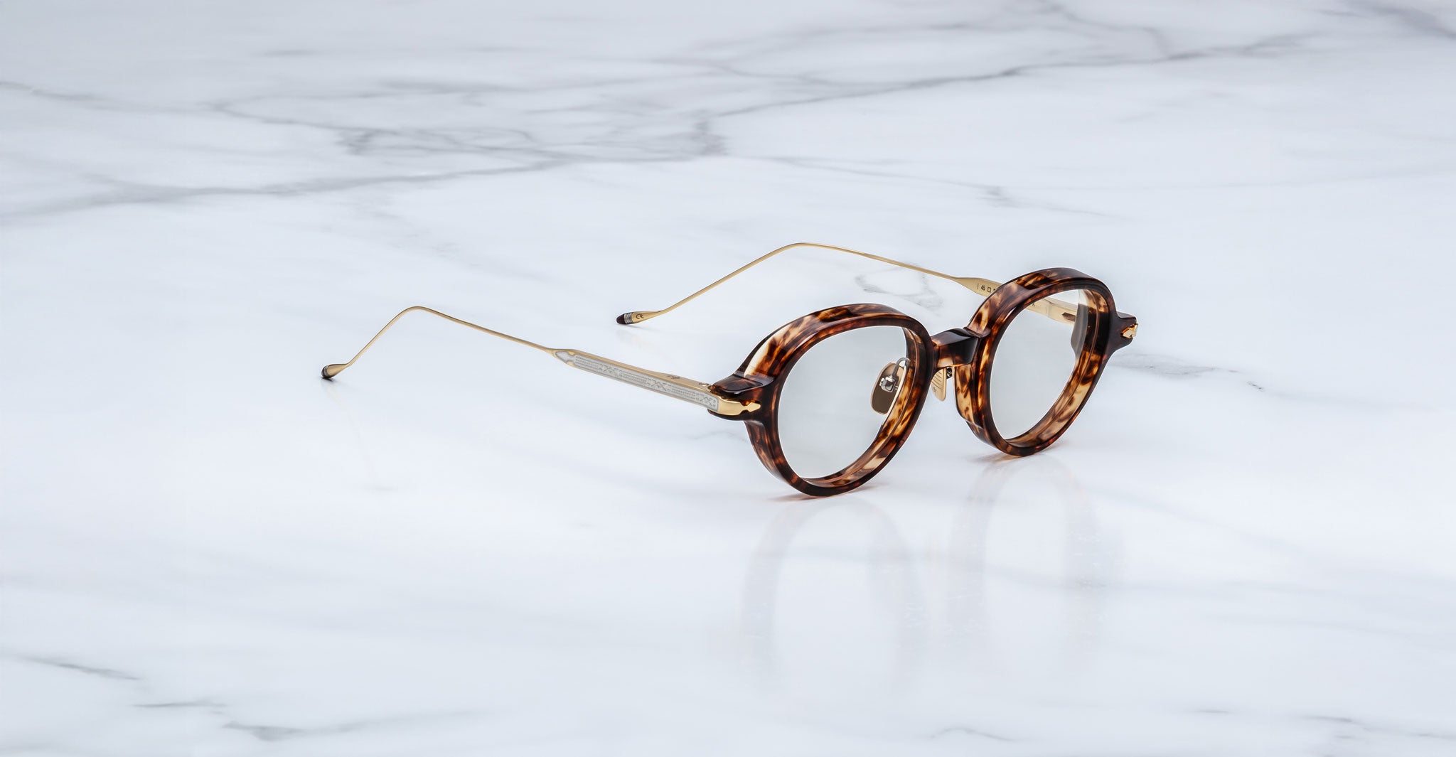 The Jacques Marie Mage Clark eyeglasses feature round tortoiseshell frames with slim gold arms, echoing midcentury style, and are shown elegantly displayed on a white marble surface.