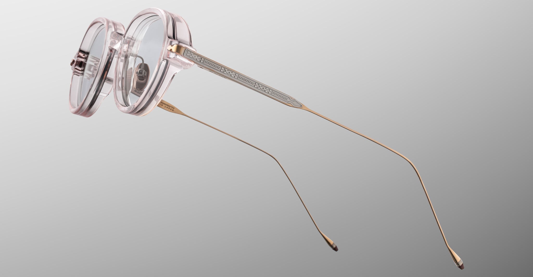 The Jacques Marie Mage Clark by Jacques Marie Mage are clear round eyeglasses with transparent pink frames and thin gold temples, inspired by midcentury design and handcrafted in Japan, shown on a light gray background.