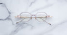 The Jacques Marie Mage Clark eyeglasses feature round, translucent frames with gold temples, reflecting midcentury style, and are displayed elegantly on a white marble surface.