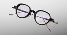 The Jacques Marie Mage Clark by Jacques Marie Mage features round, black-rimmed optical frames with thin metal arms and clear lenses, shown against a light gray background—ideal for fans of midcentury-inspired eyewear.