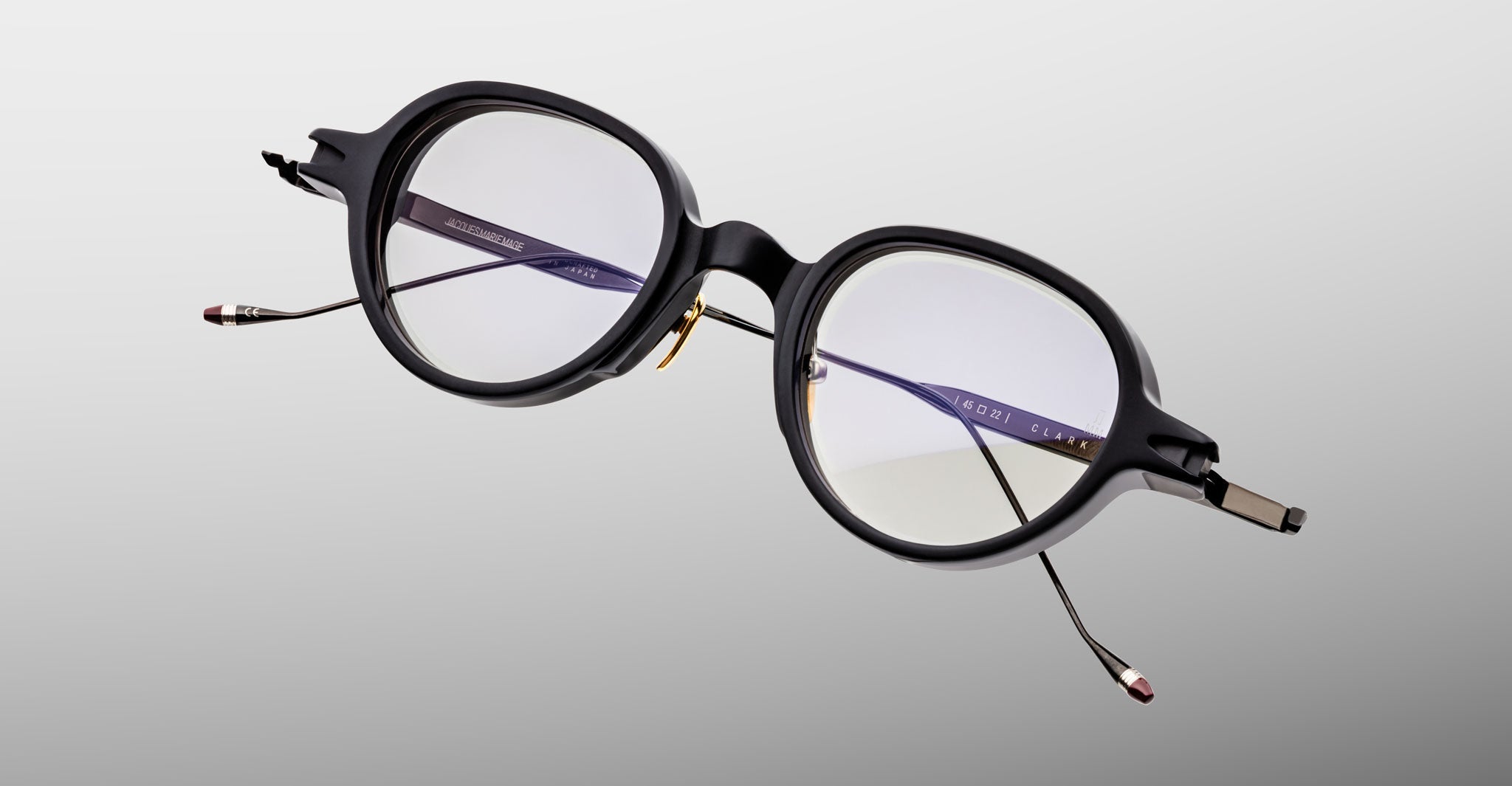 The Jacques Marie Mage Clark by Jacques Marie Mage features round, black-rimmed optical frames with thin metal arms and clear lenses, shown against a light gray background—ideal for fans of midcentury-inspired eyewear.