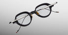 Jacques Marie Mage Clark optical frames feature black rims, silver arms, gold nose pads, and clear lenses. Expertly handcrafted in Japan, these midcentury-inspired glasses by Jacques Marie Mage appear on a light gray background.