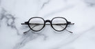 A pair of Jacques Marie Mage Clark round, black midcentury optical frames with thin temples is displayed on a white marble surface.