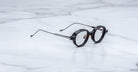 The Jacques Marie Mage Clark frames feature round black rims and thin metal arms, embodying midcentury-inspired design by Jacques Marie Mage. Shown elegantly on a white marble surface.