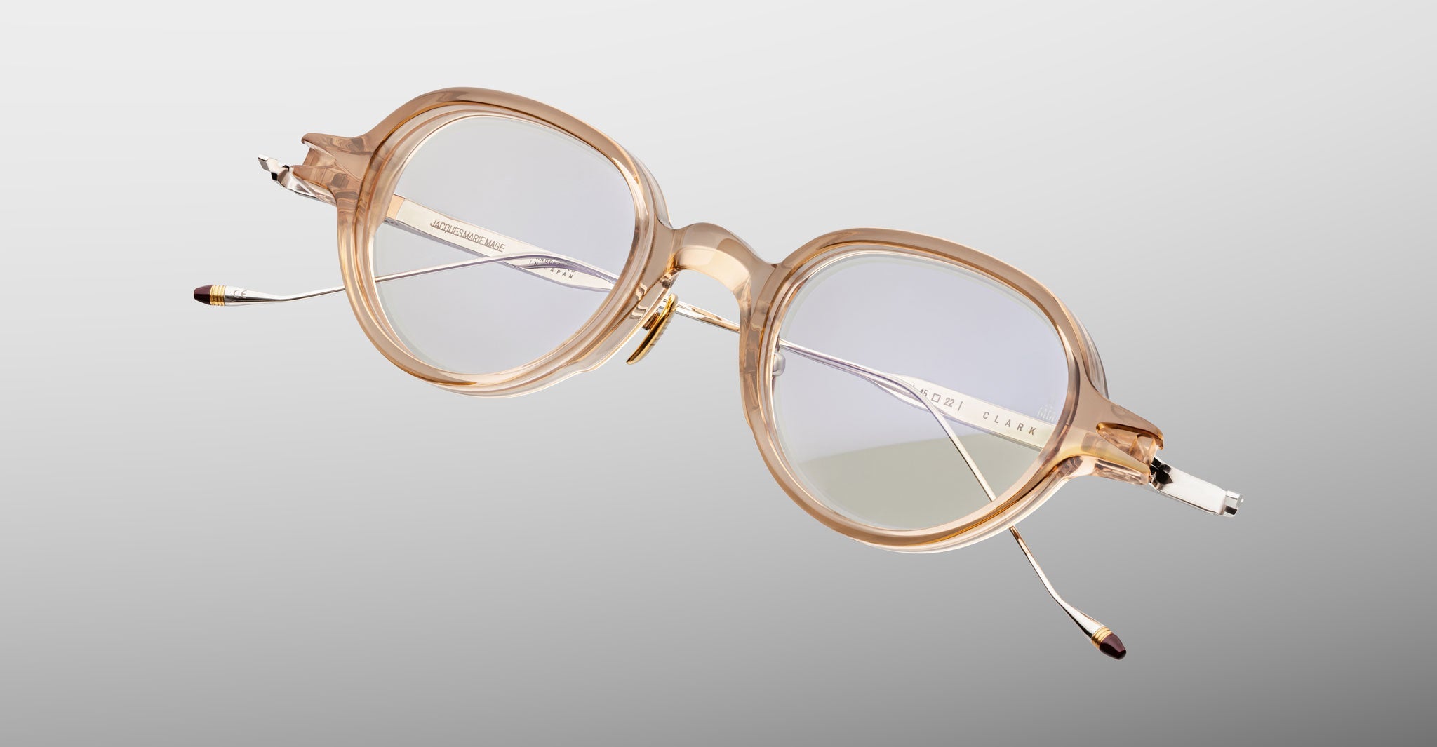 The Jacques Marie Mage Clark by Jacques Marie Mage features clear acetate, round lenses, and metal temples, inspired by mid-century optical frames and shown against a plain light gray background.