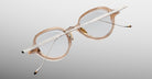 The Jacques Marie Mage Clark eyeglasses feature clear frames, metal arms, and decorative temple detailing. Handcrafted in Japan, they are displayed on a light gradient background.